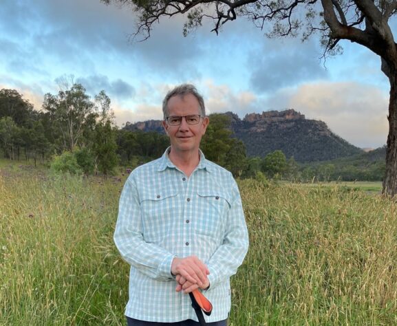 Andrew Chalk wearing a long sleeve white checked shirt with a tall large tree and towering sandstone cliffs in the far distance