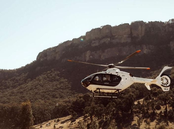 White helicopter coming into land with sandstone cliffs and steep bushland hillside in the background